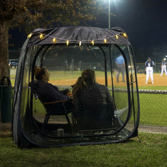 A WeatherPod pop-up tent pod in dark gray with a black detachable roof, featuring clear PVC, string lights along the top. Two women are sitting in chairs in the pod watching a baseball game at night.